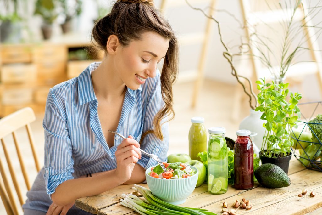 Junge und glückliche Frau, die einen gesunden Salat isst, der auf dem Tisch mit grünen frischen Zutaten drinnen sitzt