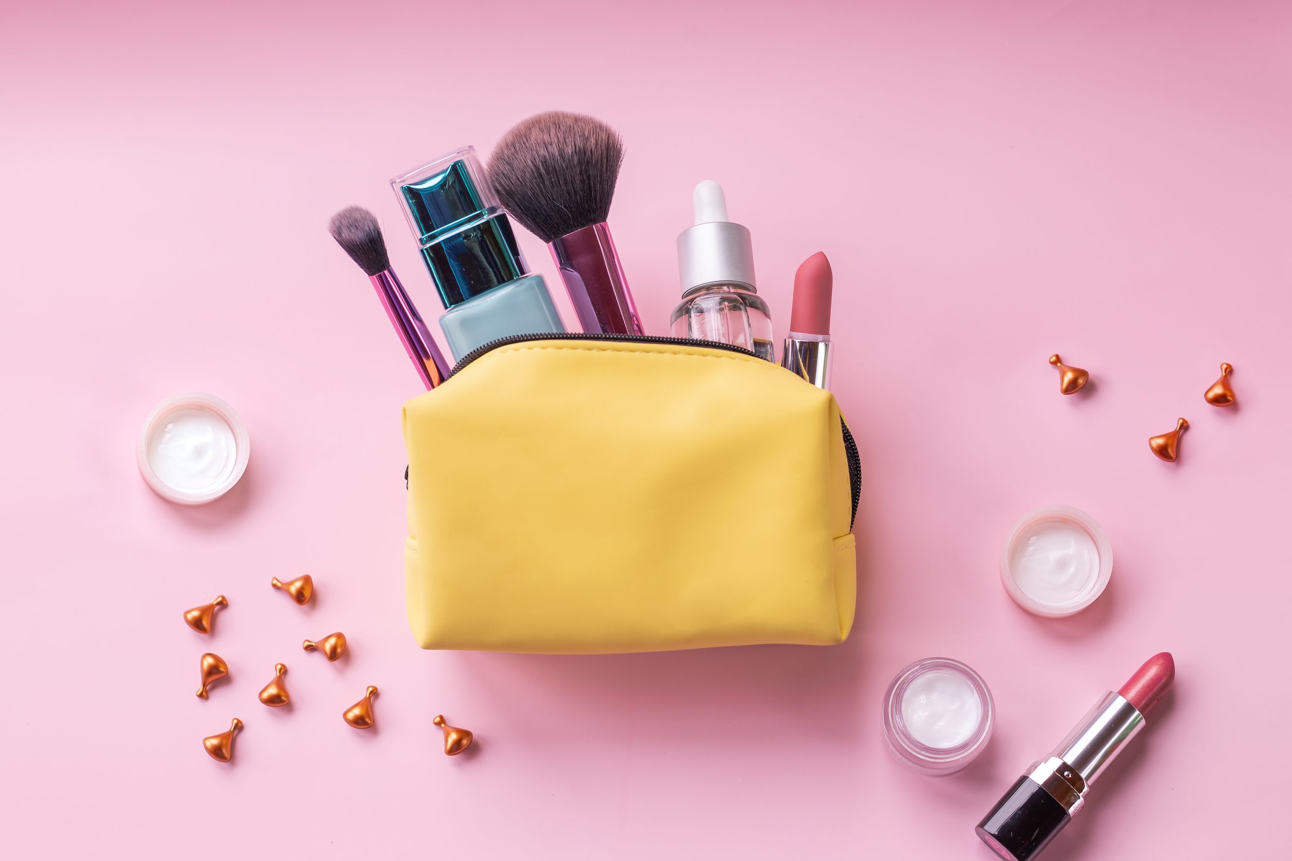 Kosmetikbeutel und Bürsten einzeln auf rosafarbenem Hintergrund. Gelbe Kosmetiktüte. Grundfarben Rosa und Gold. Hautcreme, Lippenstift, Handtasche, Kosmetikartikel