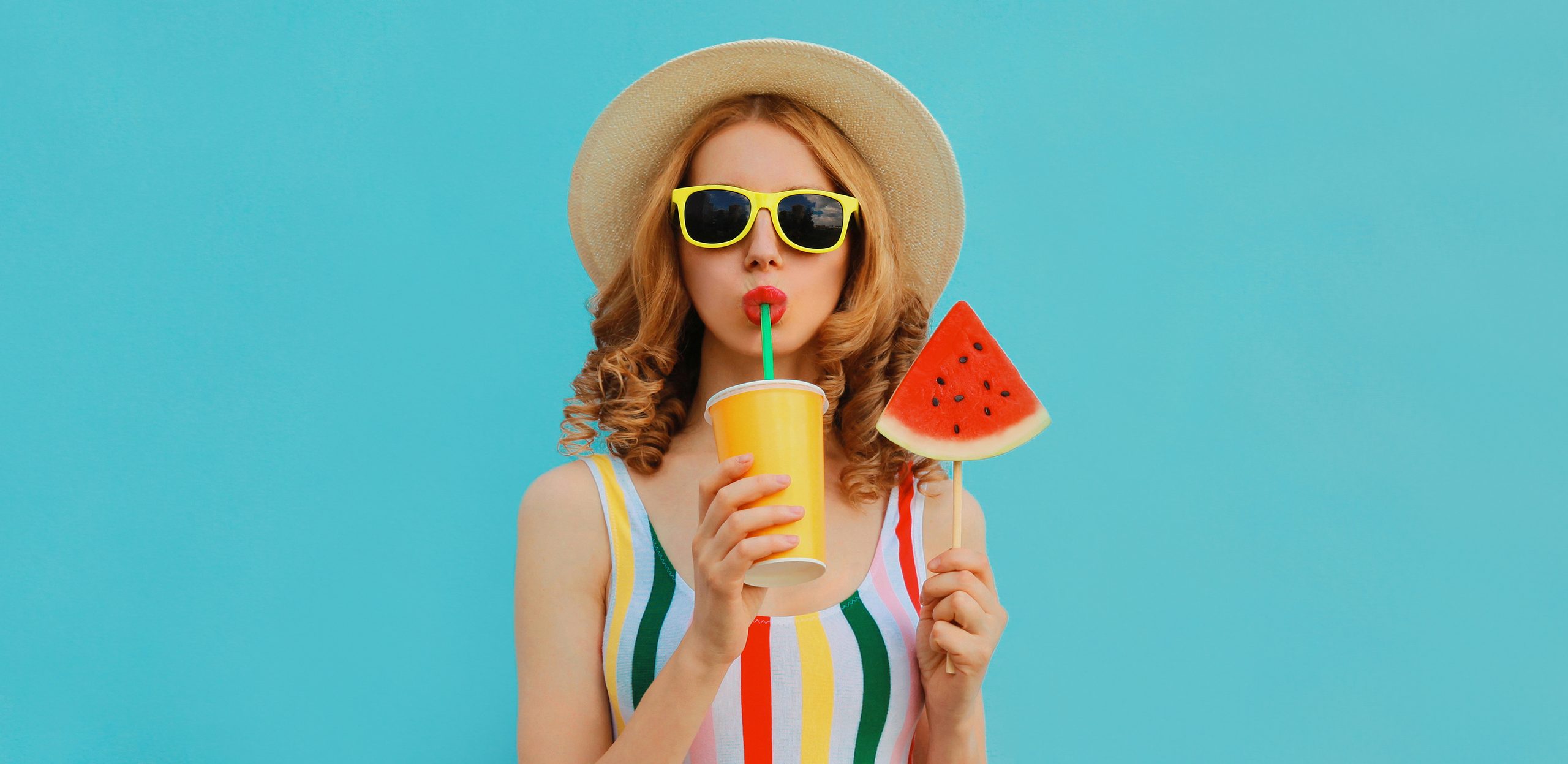 Sommerfarbiges Portrait von schöner junger Frau, die Saft mit Lollipop oder eiscremeförmigen Wassermelonen trinkt, mit einem Strohhut auf blauem Hintergrund